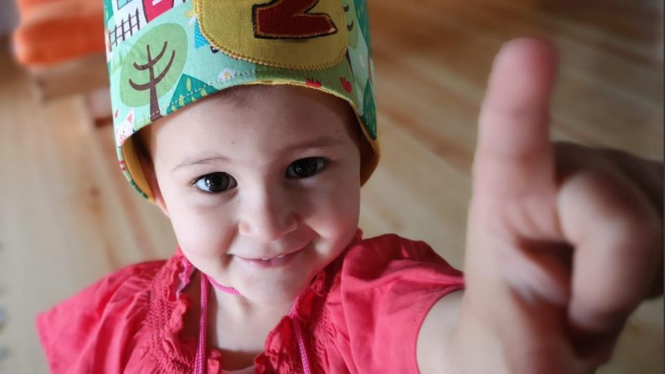 Photo taken to a 2 years-old girl who is smiling and pointing to the camera.