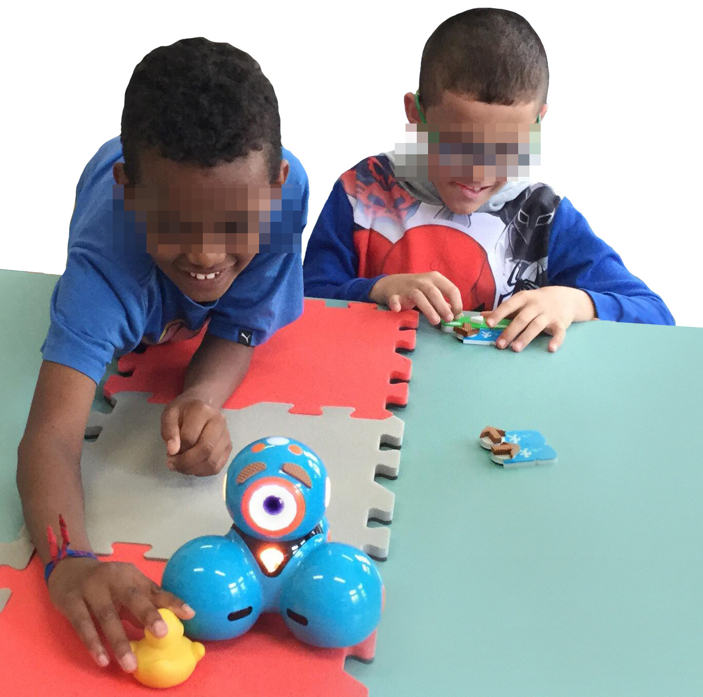 Photo taken from the Workshop with children. Two visually impaired children perform a goal-directed programming activity using a map located on the table. The activity consists of moving the robot to reach the duck. The map is made of 4 EVA foam tiles with two different colors. The child on the right is concentrated in arranging the blocks while the children at the left are touching the duck after the robot reached the duck. This child is smiling and has much of his body on the table.