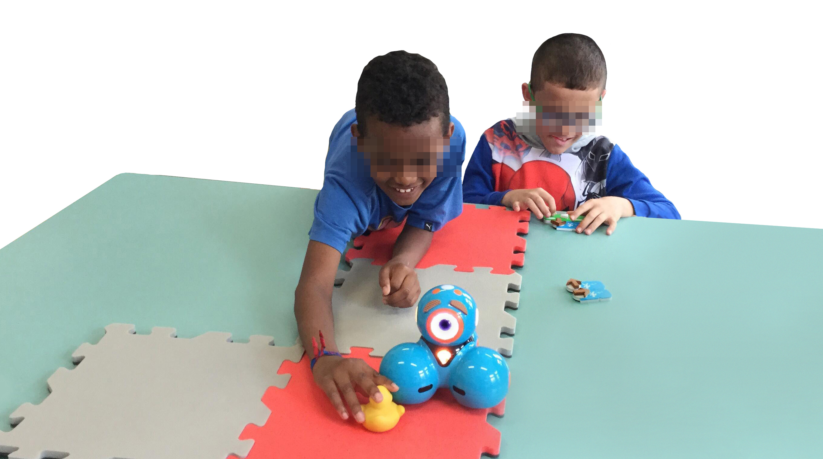 Photo taken from the Workshop with children. Two visually impaired children perform a goal-directed programming activity using a map located on the table. The activity consists of moving the robot to reach the duck. The map is made of 4 EVA foam tiles with two different colors. The child on the right is concentrated in arranging the blocks while the children at the left are touching the duck after the robot reached the duck. This child is smiling and has much of his body on the table.